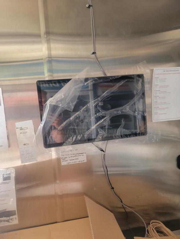 Wall-mounted monitor with protective plastic cover, visible wires, and attached notes. Appears new or unused.