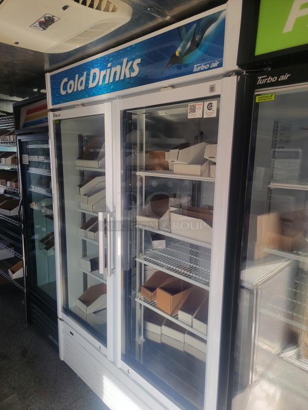 Turbo Air glass-door refrigerator for cold drinks. Features include shelves with cardboard boxes, signage, and energy-efficient label.
