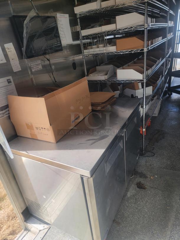 Industrial stainless steel countertop with cardboard packaging, wire shelving, and monitor. Used condition, suitable for storage or preparation.