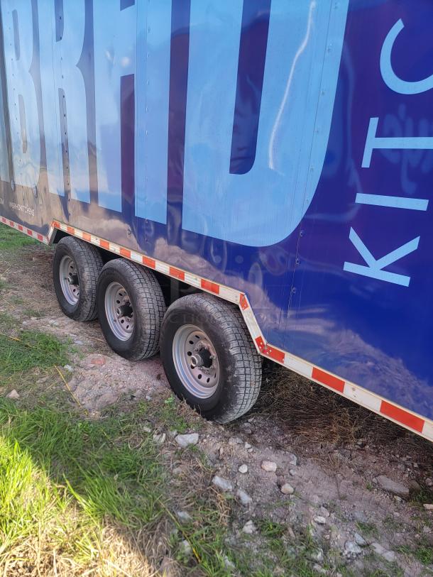 Blue triple-axle trailer with "BRAID KITCHENS" text on the side, parked on gravel.