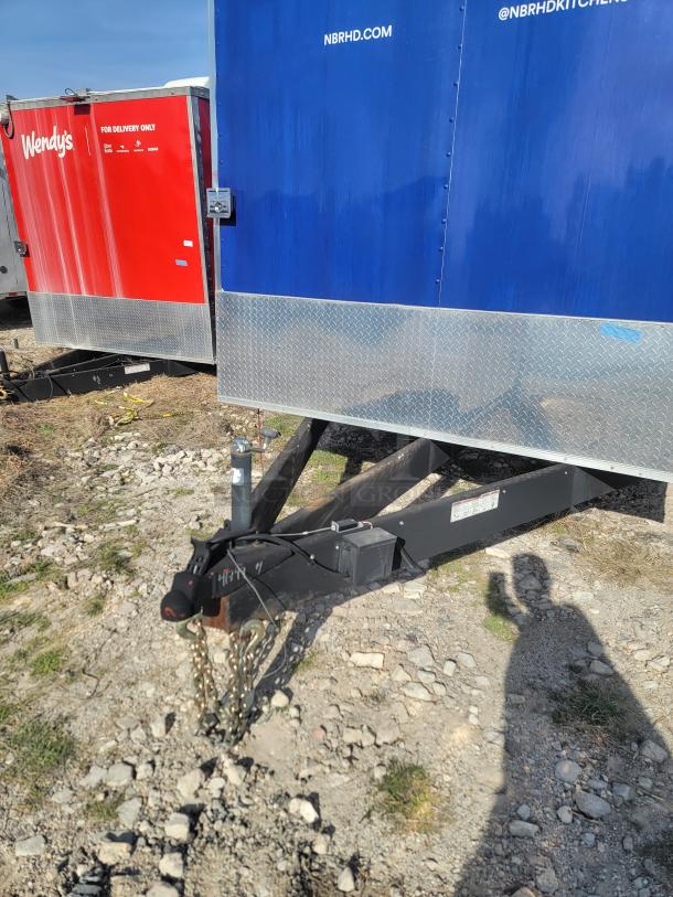 Red and blue mobile trailer for delivery use, marked with Wendy's and NBRHD branding. Visible tow hitch and chains on rocky ground.