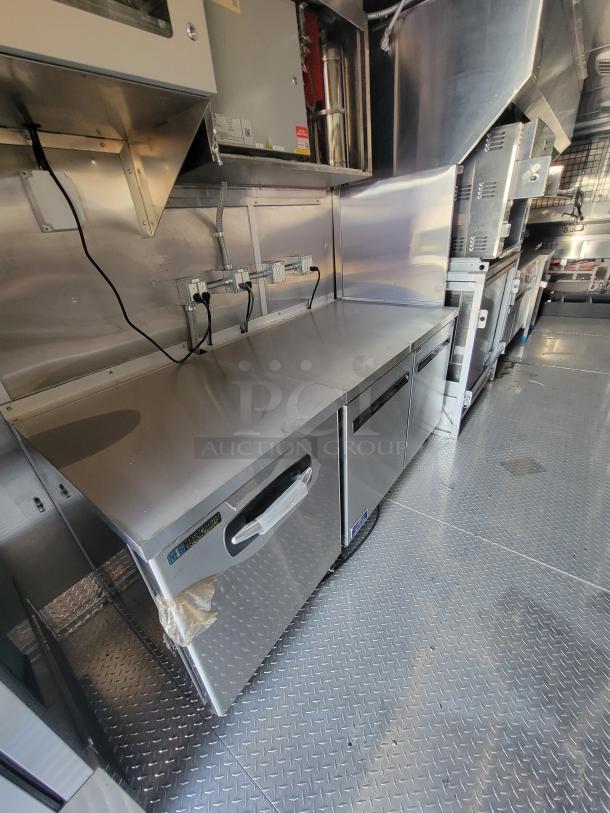 Stainless steel commercial workbench with dual refrigeration units, clean condition, visible wiring and control panel on wall.