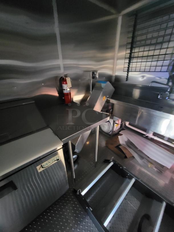 Commercial kitchen interior with stainless steel counters, Avantco refrigerator, fire extinguisher, and shelving.