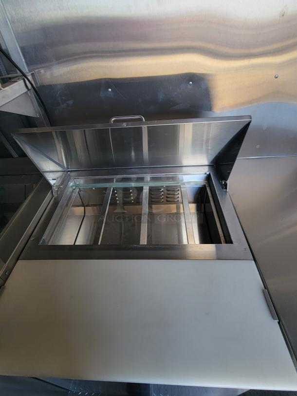 Commercial stainless steel food prep station with open lid, compartmentalized interior, and attached cutting board.