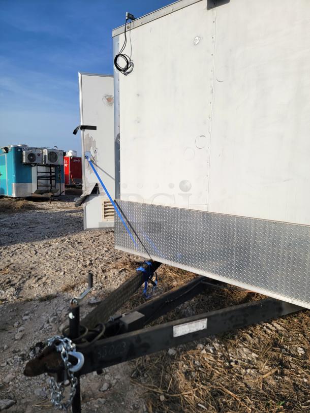 White trailer with diamond plate siding, hitched on a gravel lot. Condition appears used, with visible cables and blue ties.