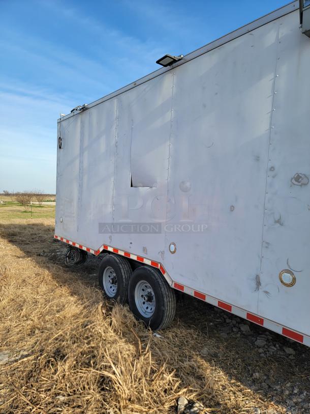 White dual-axle trailer with visible wear, dents, and a damaged panel, parked on dry grass. Ideal for restoration.