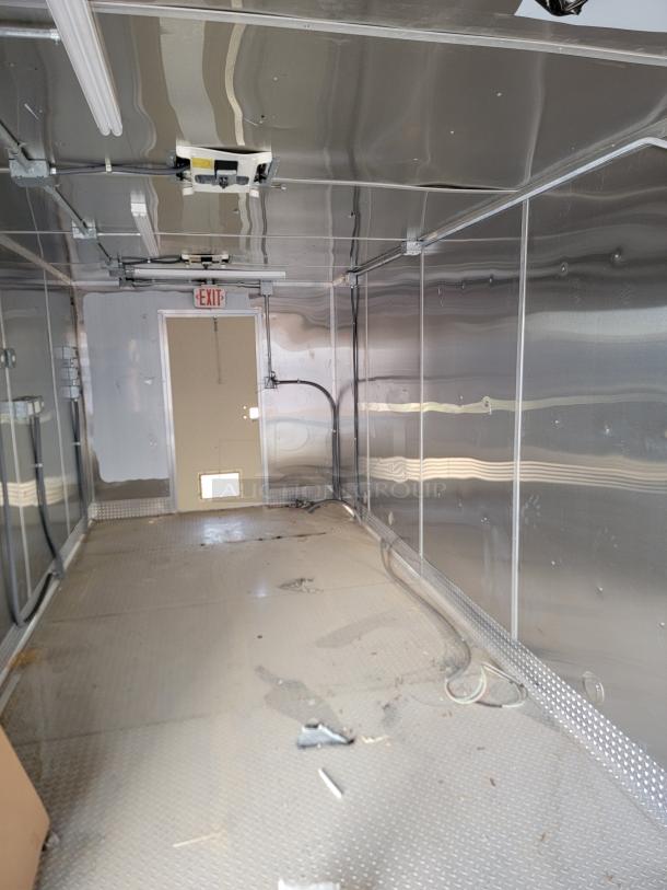 Interior of a metal cargo trailer, featuring ribbed walls and a diamond plate floor. Ceiling lights and wiring visible. Slight wear evident.