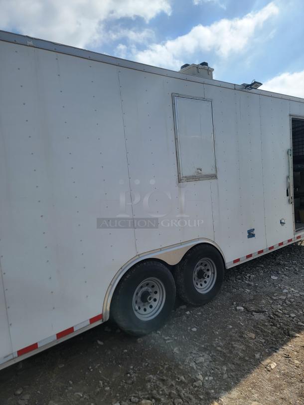 White enclosed trailer with double axles on rocky ground. Features include a side door and roof vents. In good condition.