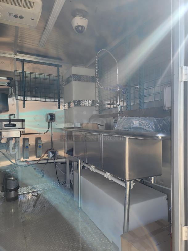 Mobile kitchen interior with stainless steel sink, plumbing, and equipment. Features security camera and air conditioning unit.