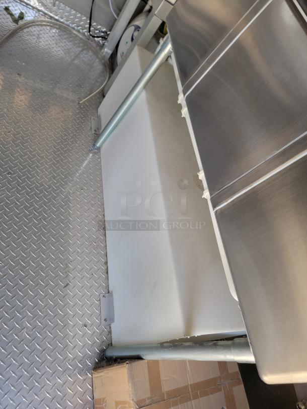 Stainless steel and white plastic equipment on diamond plate flooring, used condition. Visible support bar and hoses indicate plumbing function.
