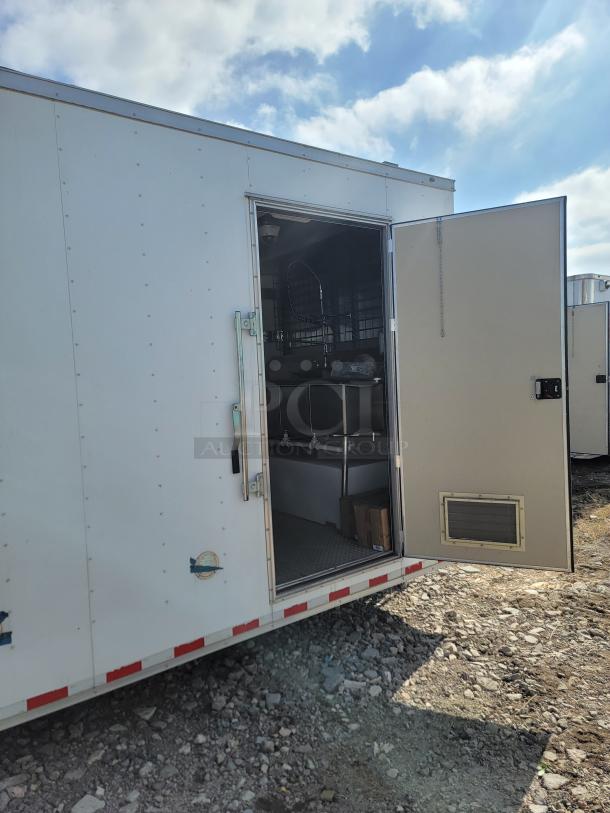 White utility trailer with an open side door, revealing interior shelving and equipment, in used condition. Rocky ground.
