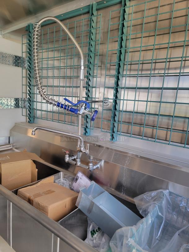 Commercial stainless steel sink with high-arc sprayer, dual-lever faucets, and various cartons in the basin. Appears unused.