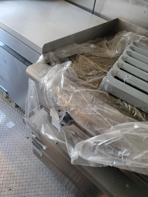 Stainless steel commercial griddle covered in plastic wrap, showing metallic grill surface and side handle. Condition appears unused.