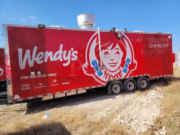 Red Wendy's food trailer with branding and delivery service logos, three axles, vent on top, parked on grass.