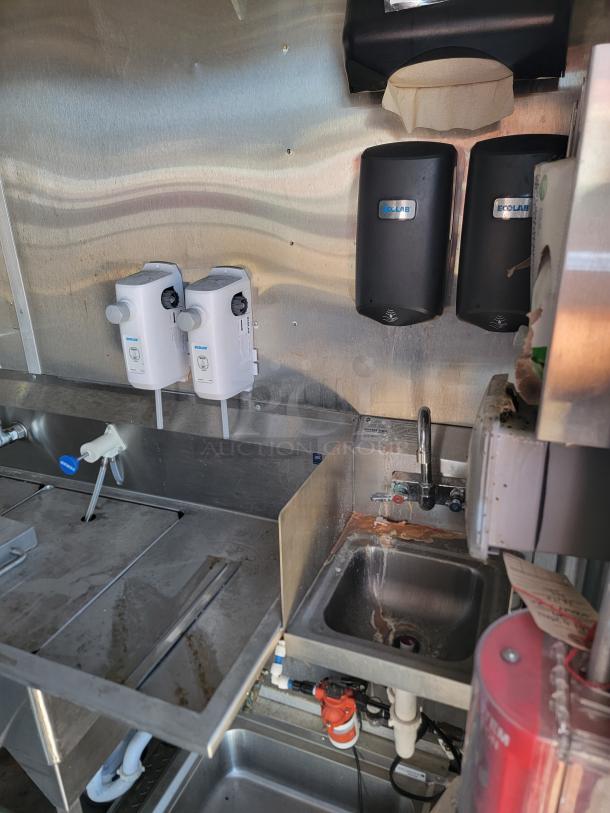 Commercial kitchen sink setup with two Ecolab dispensers, stainless steel counters, and a compact sink. Visible plumbing underneath.