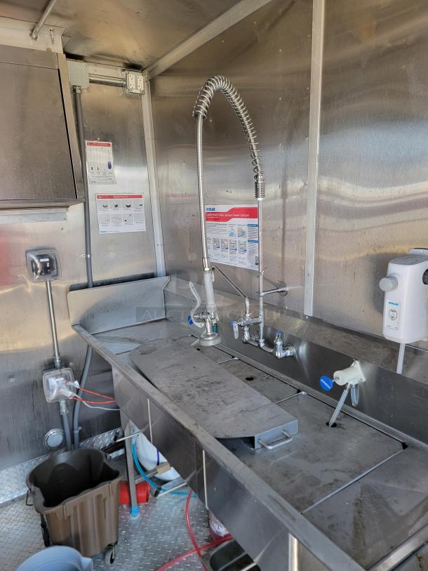 Stainless steel commercial kitchen sink with high-arch sprayer, three compartments, and various cleaning supplies. Slightly worn.