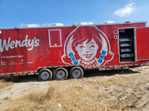 Mobile restaurant trailer with Wendy's branding, featuring a red exterior and delivery service logos. Three-axle wheels visible. Open rear door.