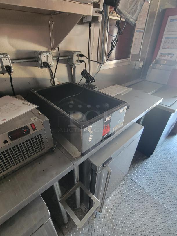 Commercial kitchen workspace with metal shelving, outlets, and under-counter equipment. Signs and papers on the counter.