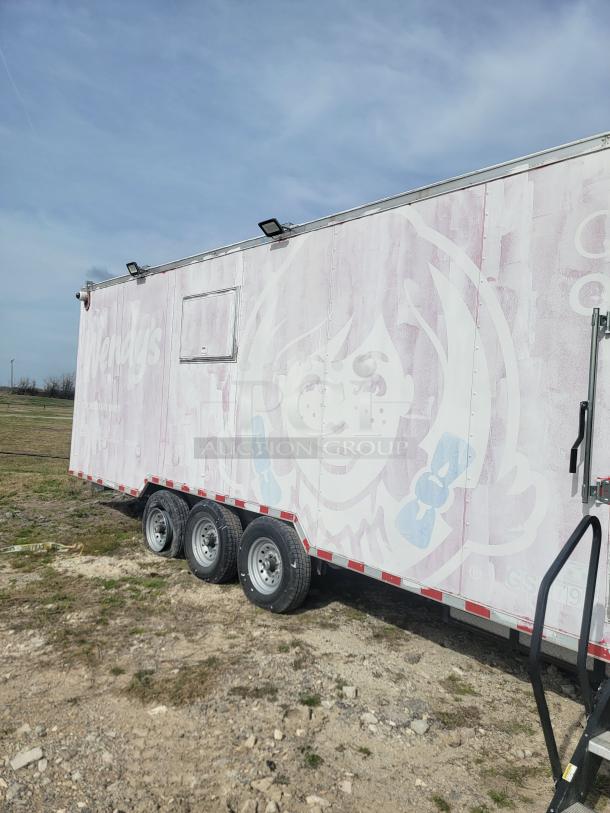 Mobile food trailer with faded branding, triple axles, and pull-down steps, situated on dirt terrain under a clear sky.