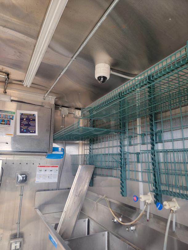 Industrial kitchen interior with metal shelves, security camera, stainless steel sink, and faucet controls.