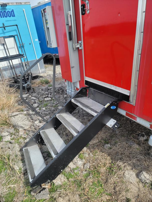 Red mobile unit with metal steps on rocky ground, featuring a black handle and visible utility boxes. Good condition.