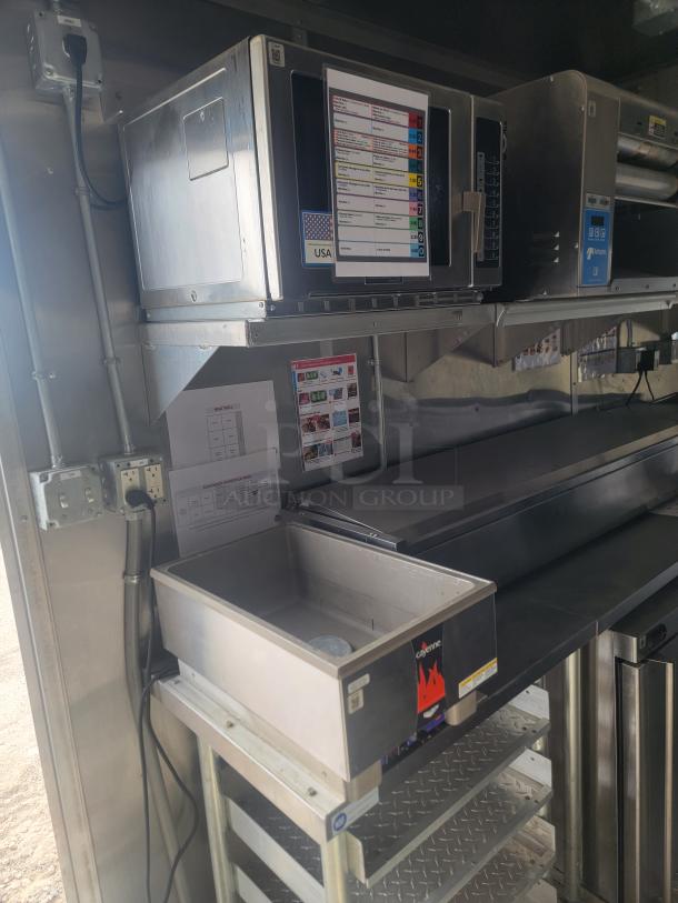 Stainless steel commercial kitchen setup with a microwave, hot food well labeled "Cayenne," and control panel. Includes plug and shelving.
