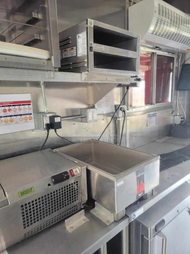 Food truck interior with stainless steel equipment including a chiller with control panel, food warmer, and HVAC unit. Visible labels indicate brand and safety instructions.