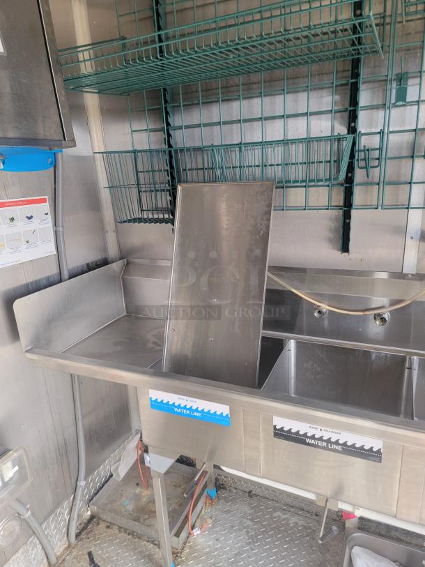 Commercial kitchen sink with dual basins and integrated shelving. Stainless steel, visible water line labels. Used condition.