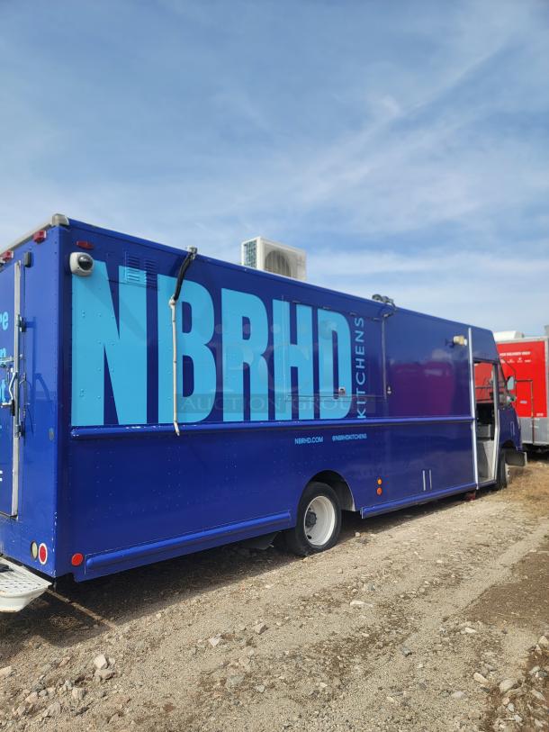 Blue food truck with "NBRHD Kitchens" text, parked outdoors. Features a flat tire and rear access step. Visible logo and website URL.