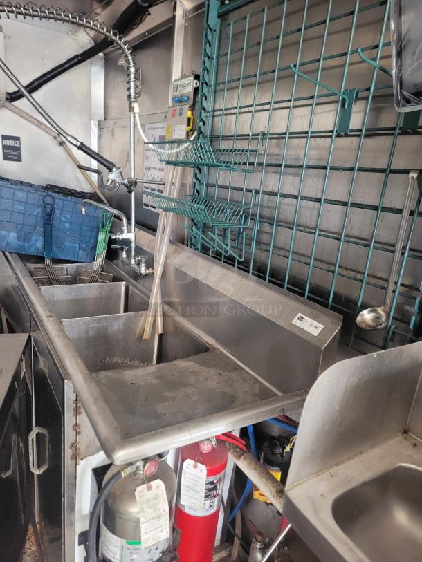 Commercial stainless steel sink with dual basins, attached spray nozzle, and mounted storage baskets. Fire extinguisher and tags visible.