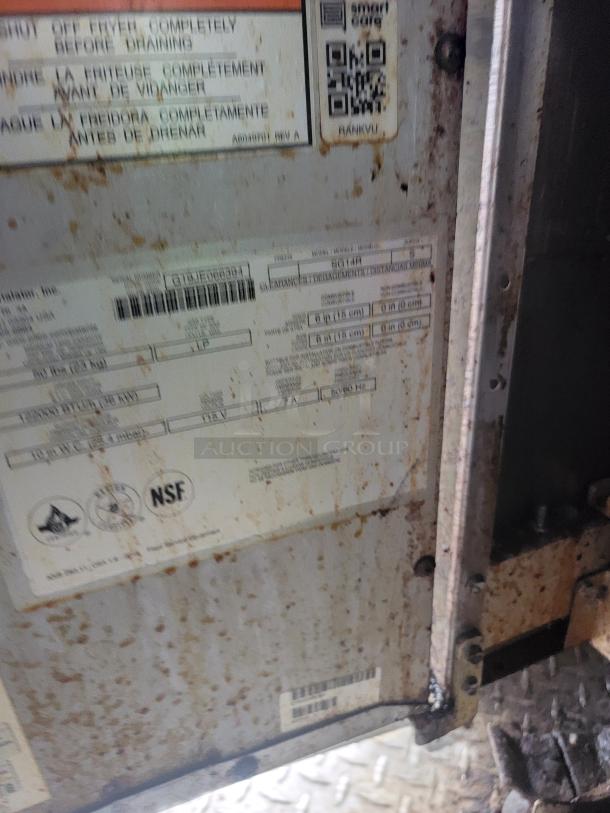 Close-up of a metal item with visible rust and label markings. Labels include QR codes, certification marks, and technical specifications.