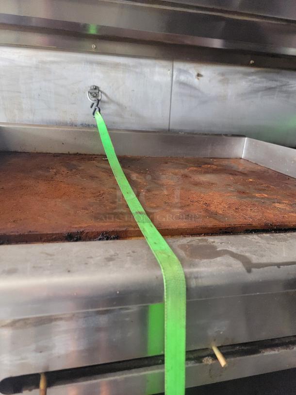 Commercial flat-top grill with a brown, rusty surface, secured with a green strap. Stainless steel body, showing signs of wear.
