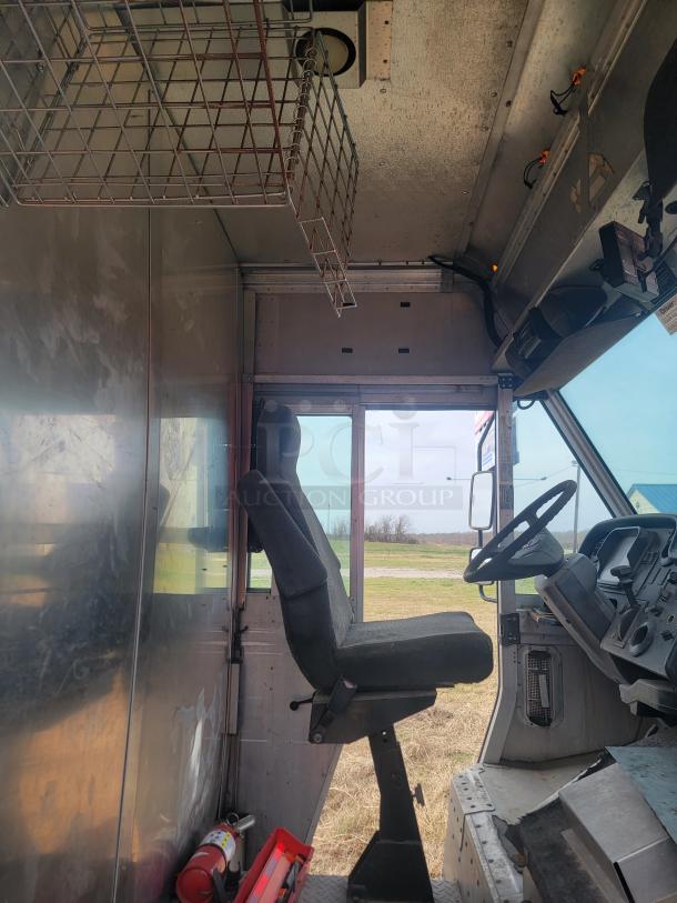 Interior view of a delivery truck cabin with driver's seat, steering wheel, and dashboard. Metal storage rack overhead and side panel. Clean condition.