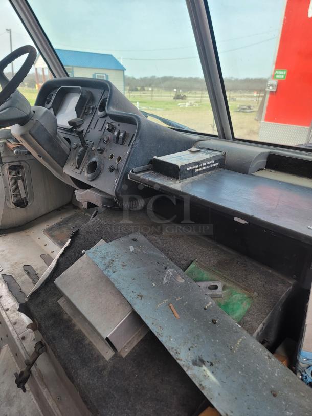 Interior view of an old vehicle dashboard with a worn, dusty control panel, steering wheel, and damaged floor carpet.