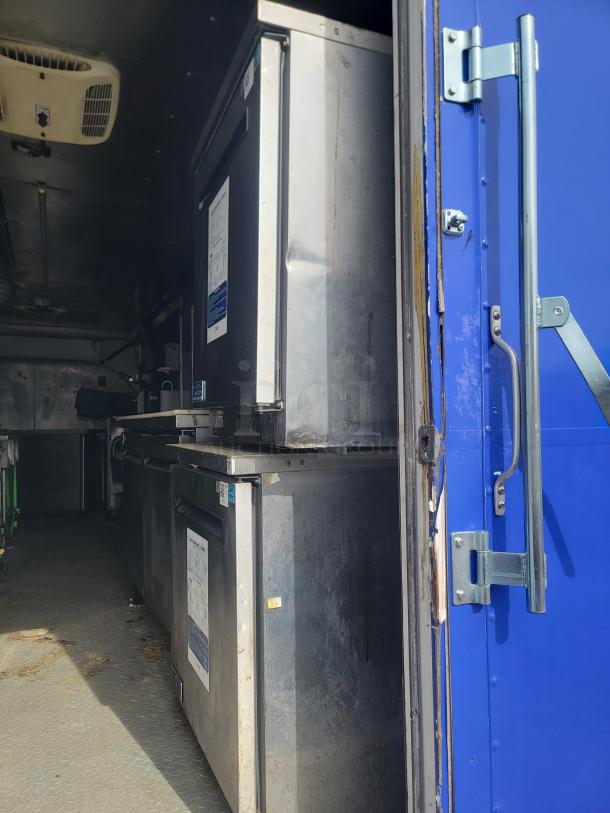 Industrial-grade stainless steel refrigerators inside a vehicle. Visible wear and printed specifications on the front. Suitable for mobile food services.