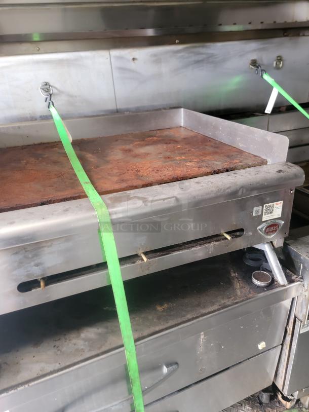 Commercial griddle with metal surface, scrapes and rust spots, secured with green straps. Label indicates "Vulcan" brand.