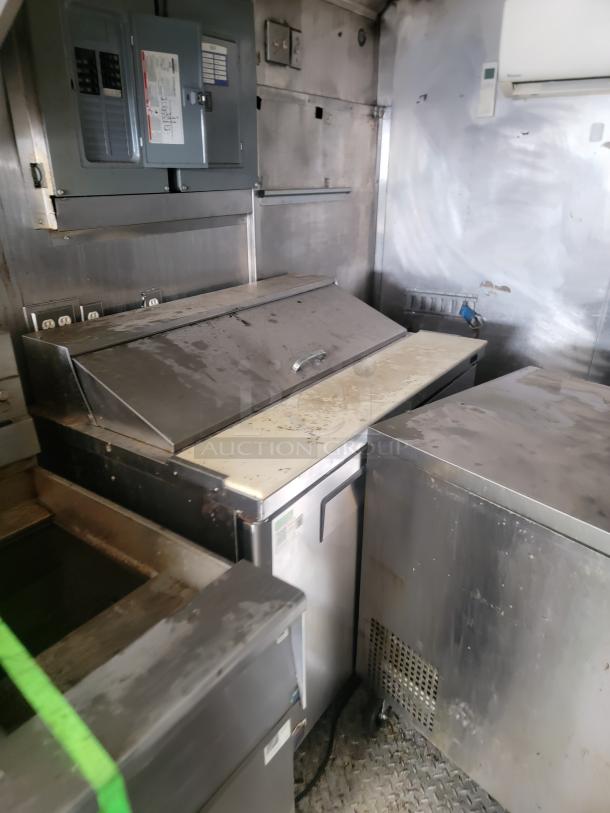 Commercial stainless steel prep table with refrigerated base. Features metal lid, electrical panel above. Worn condition.