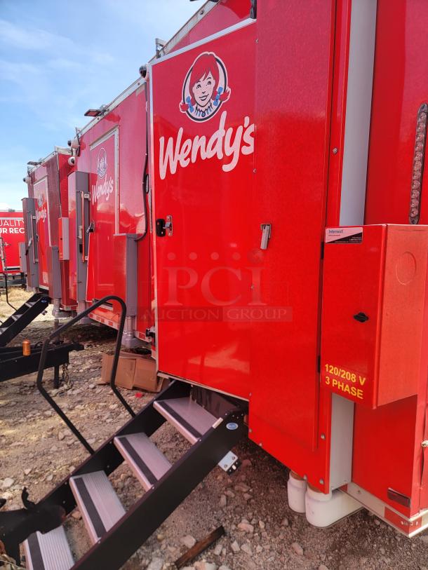 Red Wendy's food trailer with branded logo, stairs, and 120/208 V 3-phase marking, parked on rocky ground.
