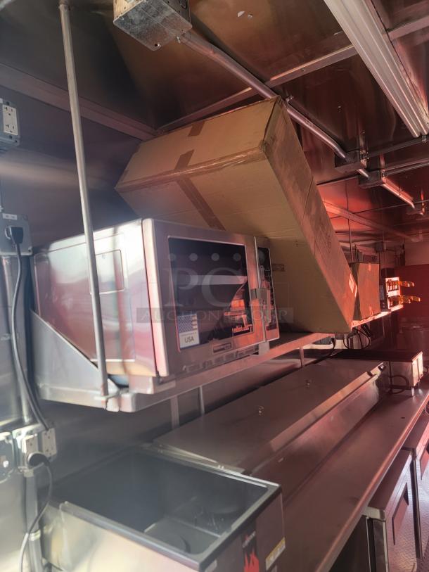 Stainless steel commercial microwave oven, model and brand unspecified, in good condition, mounted on shelf in a kitchen setup.