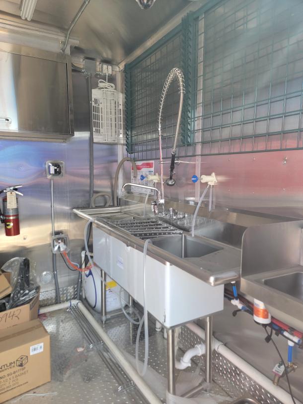 Commercial kitchen sink in stainless steel, includes dual faucets and sprayer, mounted inside a metal-walled space.