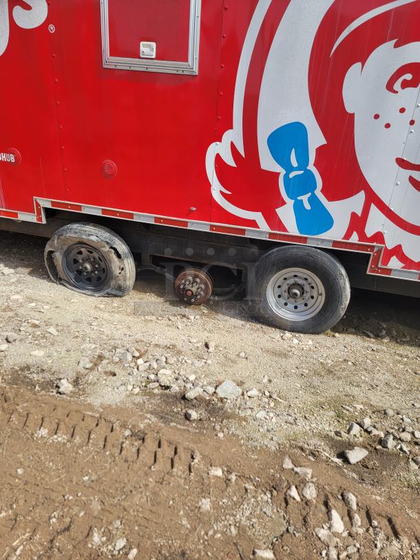 Mobile food trailer with red and white branding, missing a wheel, evident wear on remaining tires, visible serving window.