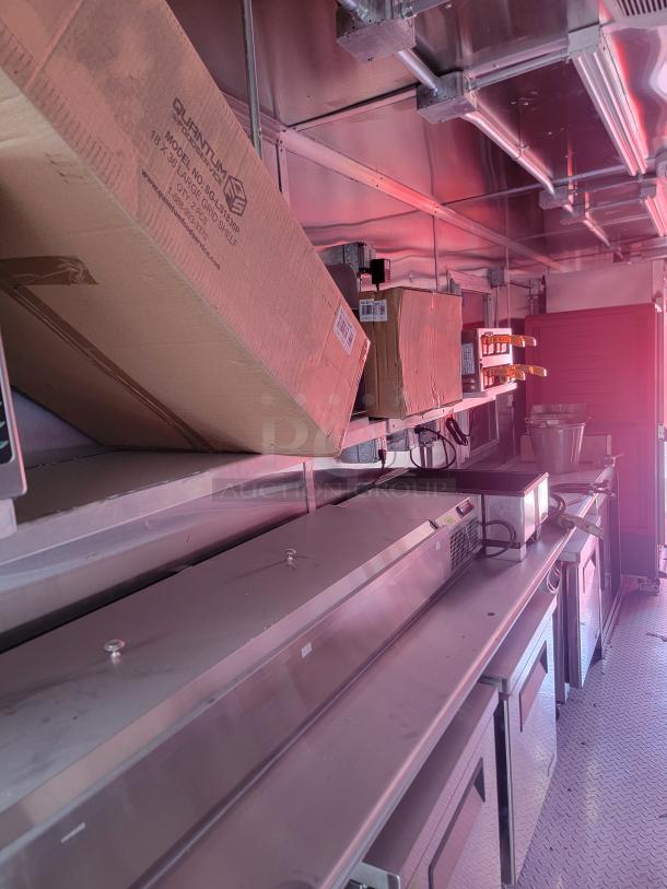 Commercial kitchen interior with stainless steel equipment, shelves, and cardboard boxes. Labels visible on boxes. Clean condition.