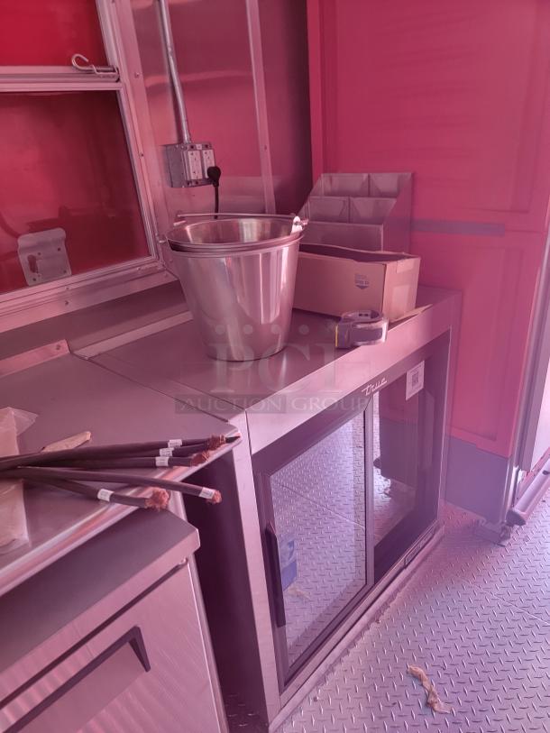 Commercial kitchen setup with a True refrigerated display unit, stainless steel bucket, and storage organizer on a metal countertop.