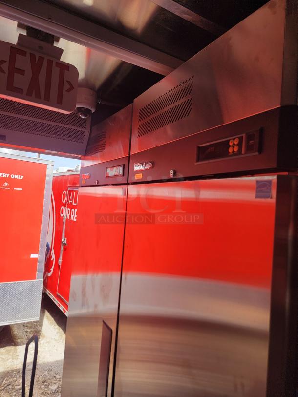 Stainless steel commercial refrigerator with digital controls, vented top, and reflective surface next to a red sign.