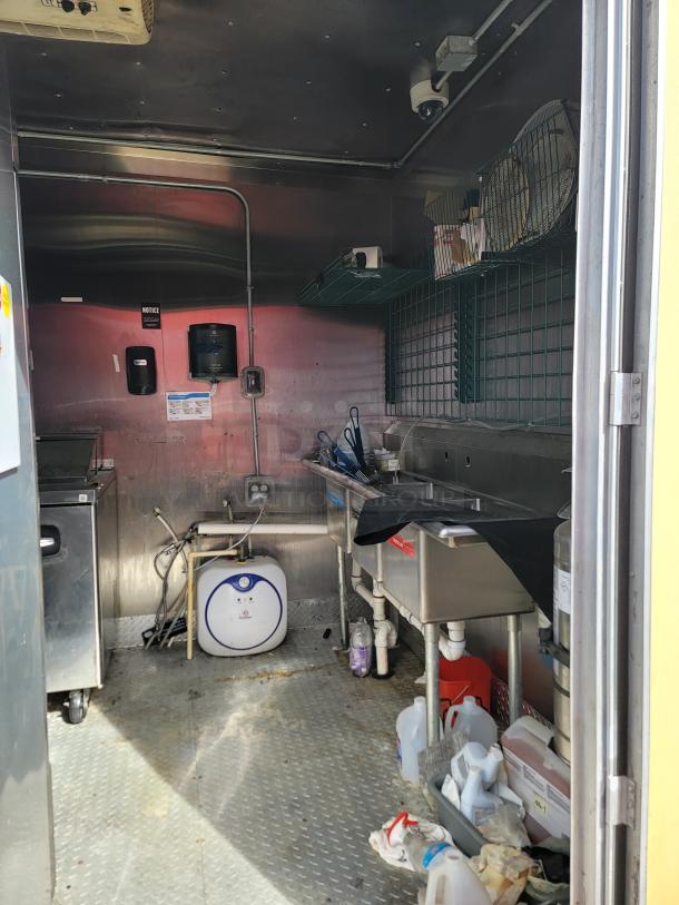 Interior of a stainless steel kitchen trailer with double sinks, fryer, shelves, storage containers, and electric connections.