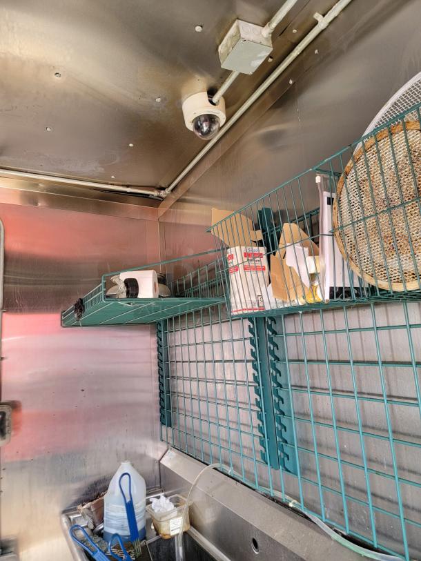 Stainless steel kitchen interior with wire shelving, surveillance camera, cleaning supplies, and a ventilation fan.