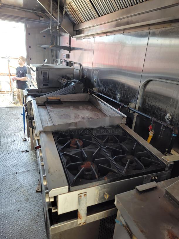 Commercial kitchen interior with a six-burner gas stove, grill, and stainless steel surfaces. Some wear visible.