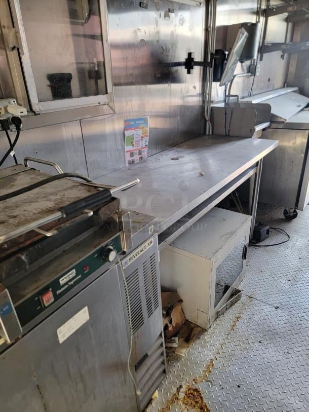 Interior of a commercial kitchen space with stainless steel counters and equipment, including a Beverage-Air refrigerator and a cooking appliance labeled "Volcano Express Steamer." The area shows signs of use and wear. A safety poster is visible on the wall.