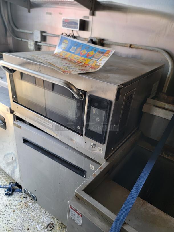 Industrial microwave oven, Amana model, stainless steel. Visible wear and grime. Features a large handle and digital controls.