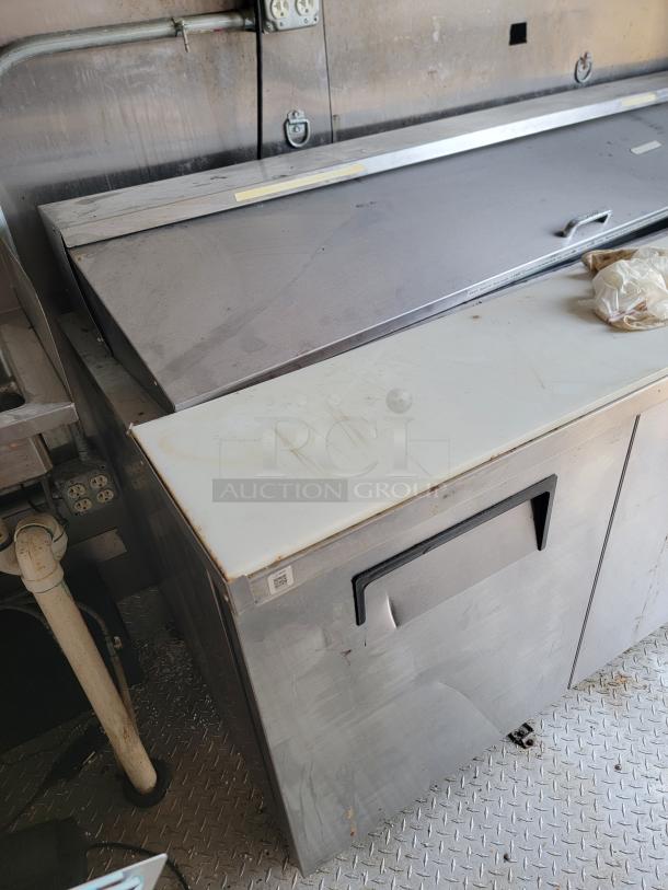 Stainless steel commercial refrigerator with white top, used condition, visible wear, features a handle and QR code tag.